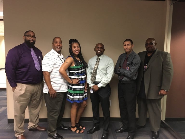 Six people standing in a group pose indoors against a beige wall, wearing business casual and formal attire