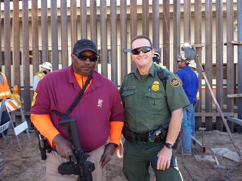 Two men smiling at camera, one in purple and orange shirt with cap, one in green uniform with badge, standing in front of metal fence