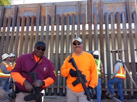 Two workers in safety vests and hard hats holding power tools stand in front of an industrial metal fence